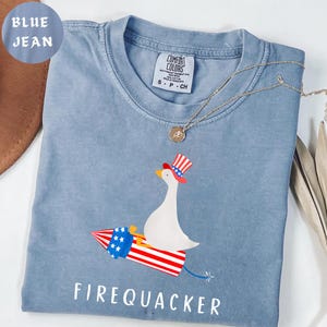 May include: Blue jean colored t-shirt with a graphic of a white goose wearing a patriotic hat, riding a red, white, and blue firework. The text "FIREQUACKER" is printed below the graphic. A gold necklace is draped across the shirt.