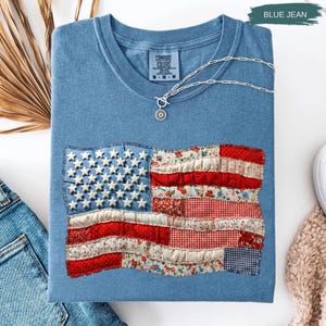 May include: A blue jean colored t-shirt with a sewn-on American flag design. The flag features white stars on a blue field and red and white stripes made of various patterned fabrics. A silver necklace with a pendant rests on the shirt.
