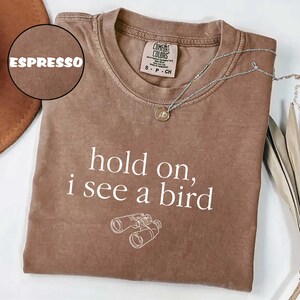 May include: Espresso-colored t-shirt with the text "hold on, i see a bird" and a graphic of binoculars. The shirt has a crew neck and short sleeves. A silver necklace with a pendant is draped across the shirt.