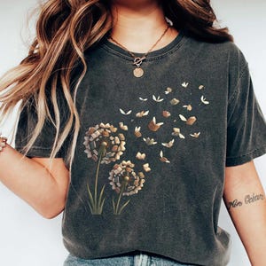 May include: A charcoal gray short-sleeve t-shirt featuring a graphic print of two dandelion-like flowers where the seeds are replaced by small open books. The books appear to be floating away from the stems, creating a whimsical, airy effect.