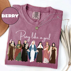 May include: Berry-colored t-shirt with the phrase "Pray like a girl" in cursive. The shirt features an illustration of seven women in religious attire. A silver necklace with a pendant is draped across the shirt.