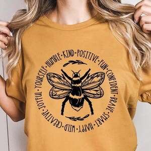 May include: A mustard yellow t-shirt with a black bee graphic and a circle of words around it. The words are: "Smart, Brave, Confident, Fun, Positive, Kind, Humble, Yourself, Joyful, Creative, Wild, Happy."