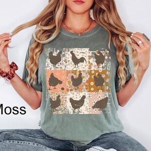 May include: A moss green t-shirt featuring a patchwork design with brown chicken silhouettes on floral and solid color backgrounds. The word "Moss" is printed in black on the left side of the shirt.