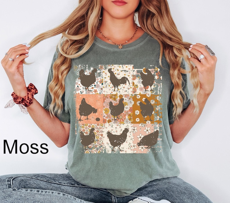 May include: A moss green t-shirt featuring a patchwork design with brown chicken silhouettes on floral and solid color backgrounds. The word "Moss" is printed in black on the left side of the shirt.