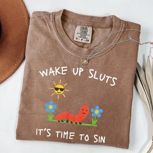 Puede incluir: Camiseta Comfort Colors marr&oacute;n con el texto "WAKE UP SLUTS IT'S TIME TO SIN". El gr&aacute;fico incluye un sol sonriente con gafas de sol, un gusano rojo y flores azules.