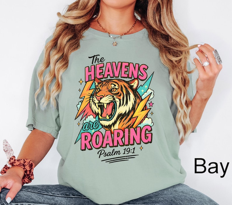 Puede incluir: Camiseta verde salvia con un gr&aacute;fico colorido de un tigre rugiendo, rel&aacute;mpagos y el texto "The Heavens are Roaring" en rosa y blanco. Incluye el vers&iacute;culo b&iacute;blico Salmo 19:1.