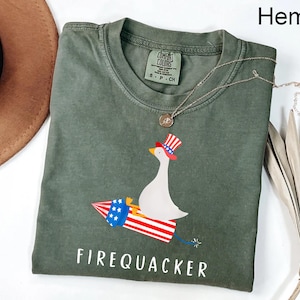 K&ouml;nnte beinhalten: Ein hanffarbenes Kurzarm-T-Shirt mit einer Grafik einer wei&szlig;en Gans, die einen rot-wei&szlig;-blauen Zylinder tr&auml;gt und auf einer Feuerwerksrakete reitet. Der Text FIREQUACKER ist darunter in wei&szlig;en Gro&szlig;buchstaben aufgedruckt.