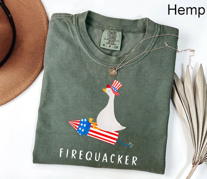 K&ouml;nnte beinhalten: Ein hanffarbenes Kurzarm-T-Shirt mit einer Grafik einer wei&szlig;en Gans, die einen rot-wei&szlig;-blauen Zylinder tr&auml;gt und auf einer Feuerwerksrakete reitet. Der Text FIREQUACKER ist darunter in wei&szlig;en Gro&szlig;buchstaben aufgedruckt.