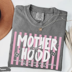 May include: A gray t-shirt with the word "MOTHERHOOD" in white, accented with pink vertical stripes. The shirt includes the text "some days I rock it some days it rocks me either way we're rockin'". A silver necklace with a pendant is on the shirt.
