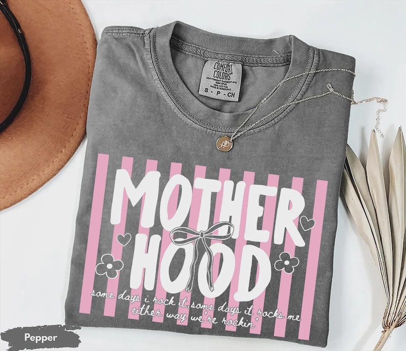 May include: A gray t-shirt with the word "MOTHERHOOD" in white, accented with pink vertical stripes. The shirt includes the text "some days I rock it some days it rocks me either way we're rockin'". A silver necklace with a pendant is on the shirt.