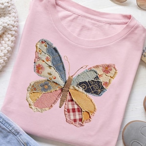 May include: A light pink t-shirt featuring a patchwork butterfly design. The butterfly's wings are made of various patterned fabrics in shades of blue, pink, yellow, and red. The shirt has a crew neck and short sleeves, perfect for casual wear.
