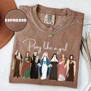 May include: Espresso-colored t-shirt with the text "Pray like a girl" and an illustration of seven figures in religious attire. A silver necklace with a pendant rests on the shirt. The shirt is a Comfort Colors brand.