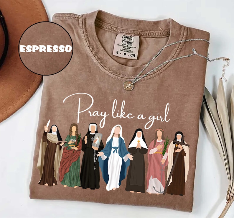 May include: Espresso-colored t-shirt with the text "Pray like a girl" and an illustration of seven figures in religious attire. A silver necklace with a pendant rests on the shirt. The shirt is a Comfort Colors brand.
