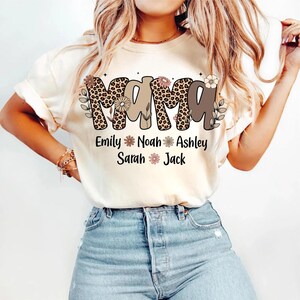 May include: Cream-colored t-shirt with the word "MAMA" in a leopard print design and brown letters. Below the word "MAMA" are the names Emily, Noah, Ashley, Sarah, and Jack. Floral accents are included in the design.