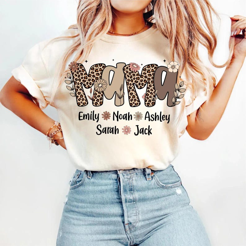 May include: Cream-colored t-shirt with the word "MAMA" in a leopard print design and brown letters. Below the word "MAMA" are the names Emily, Noah, Ashley, Sarah, and Jack. Floral accents are included in the design.