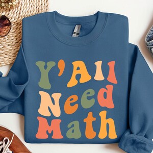 K&ouml;nnte beinhalten: Ein blaues Sweatshirt mit dem Text "Y'all Need Math" in bunten, Retro-Lettern.