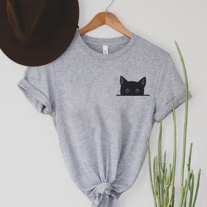 May include: A heather gray short-sleeved t-shirt featuring a black graphic of a cat head peeking over a horizontal line on the left chest area. The shirt has a crew neckline and rolled sleeves, displayed on a wooden hanger with a brown hat.