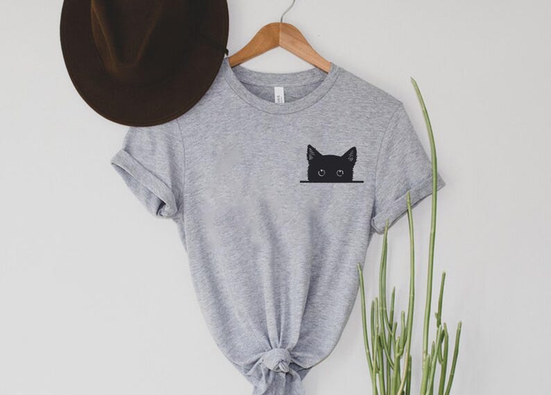 May include: A heather gray short-sleeved t-shirt featuring a black graphic of a cat head peeking over a horizontal line on the left chest area. The shirt has a crew neckline and rolled sleeves, displayed on a wooden hanger with a brown hat.
