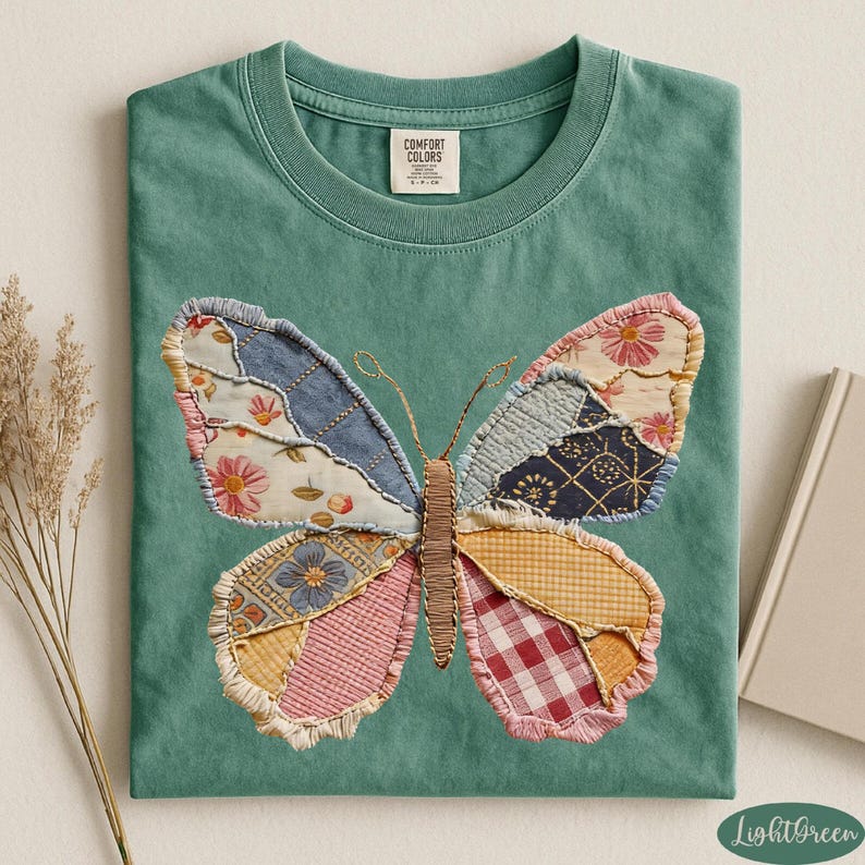 May include: A light green t-shirt featuring a large butterfly design. The butterfly is made of various fabric patches in shades of blue, pink, yellow, and red. The shirt has a crew neck and short sleeves. The brand label "Comfort Colors" is visible.