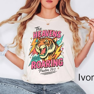 Puede incluir: Camiseta color marfil con una ilustraci&oacute;n de tigre y el texto "The Heavens are Roaring" en rosa y azul. El dise&ntilde;o incluye rayos y el vers&iacute;culo b&iacute;blico Salmo 19:1.