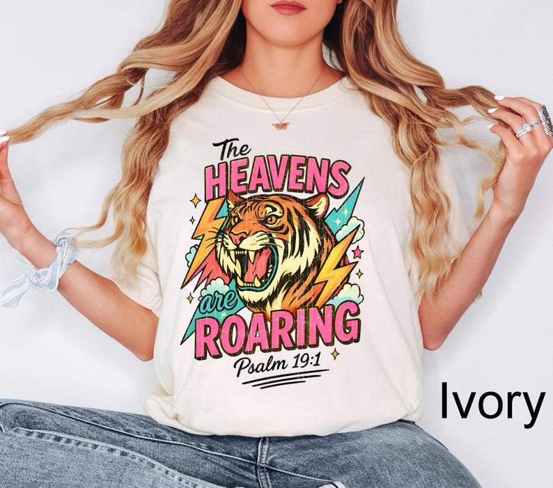 Puede incluir: Camiseta color marfil con una ilustraci&oacute;n de tigre y el texto "The Heavens are Roaring" en rosa y azul. El dise&ntilde;o incluye rayos y el vers&iacute;culo b&iacute;blico Salmo 19:1.