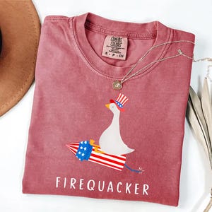 K&ouml;nnte beinhalten: Ein altrosa T-Shirt mit einer Grafik einer wei&szlig;en Gans, die einen rot-wei&szlig;-blau gestreiften Zylinder tr&auml;gt und auf einer Feuerwerksrakete in passenden patriotischen Farben reitet. Der Text FIREQUACKER ist darunter in Wei&szlig; gedruckt.