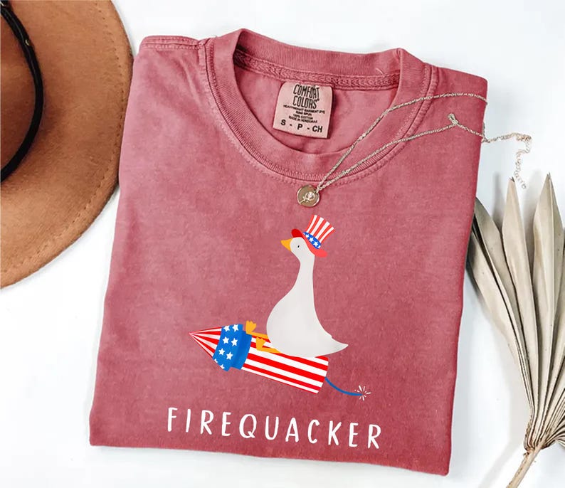 K&ouml;nnte beinhalten: Ein altrosa T-Shirt mit einer Grafik einer wei&szlig;en Gans, die einen rot-wei&szlig;-blau gestreiften Zylinder tr&auml;gt und auf einer Feuerwerksrakete in passenden patriotischen Farben reitet. Der Text FIREQUACKER ist darunter in Wei&szlig; gedruckt.