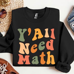 K&ouml;nnte beinhalten: Schwarzer Pullover mit dem Text "Y'all Need Math" in bunter Retro-Schrift.