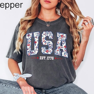May include: A dark gray t-shirt with the word "USA" in large, floral-patterned letters. The shirt also features the text "EST. 1776" in red. The shirt is a casual style, suitable for everyday wear.