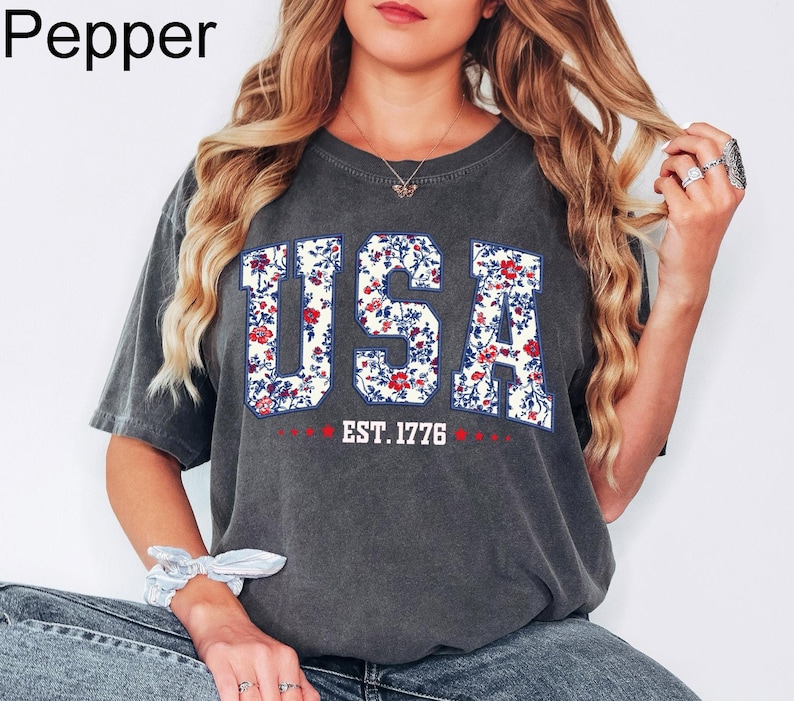 May include: A dark gray t-shirt with the word "USA" in large, floral-patterned letters. The shirt also features the text "EST. 1776" in red. The shirt is a casual style, suitable for everyday wear.