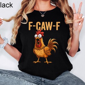 May include: A black short-sleeved t-shirt featuring a graphic of a wide-eyed, surprised-looking brown rooster with a red comb and wattle. Above the rooster, the text F-CAW-F is printed in a bold, light-colored, sans-serif font.