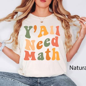 K&ouml;nnte beinhalten: Ein wei&szlig;es T-Shirt mit dem Text "Y'all Need Math" in bunten Retro-Lettern. Das Shirt wird von einer Person mit langen blonden Haaren modelliert. Der Hintergrund ist eine schlichte wei&szlig;e Wand. Der Text "Natur" ist in der rechten unteren Ecke des Bildes sichtbar.