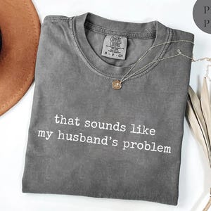 May include: A grey Comfort Colors t-shirt with the slogan "that sounds like my husband's problem" in white. A silver necklace with a circular pendant is on the shirt. A brown hat and dried leaves are in the background.
