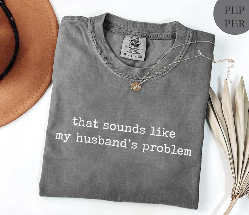 May include: A grey Comfort Colors t-shirt with the slogan "that sounds like my husband's problem" in white. A silver necklace with a circular pendant is on the shirt. A brown hat and dried leaves are in the background.
