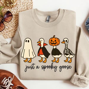 以下が含まれることがあります： ハロウィーンをテーマにしたベージュのスウェットシャツ。デザインは、幽霊、吸血鬼、カボチャ、スケルトンに扮した4羽の漫画のガチョウが特徴です。「just a spooky goose」というテキストがガチョウの下にあります。スウェットシャツはジーンズと茶色の靴と組み合わせています。