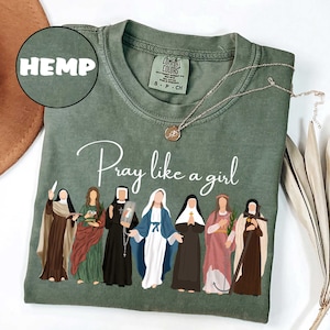 May include: Sage green t-shirt with the words "Pray like a girl" and an illustration of several women. The shirt also has the word "HEMP" in a circle. A gold necklace with a pendant is on the shirt.