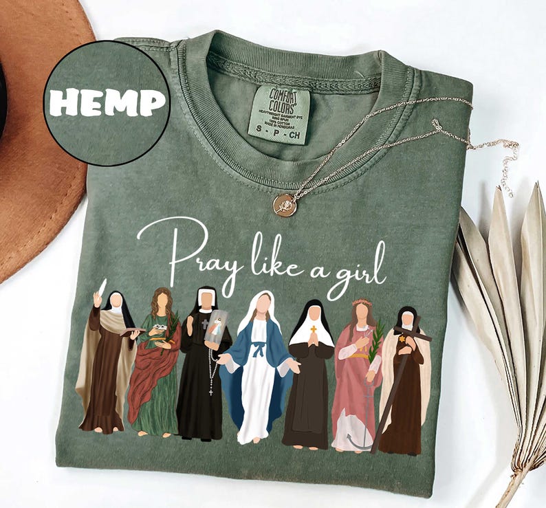 May include: Sage green t-shirt with the words "Pray like a girl" and an illustration of several women. The shirt also has the word "HEMP" in a circle. A gold necklace with a pendant is on the shirt.