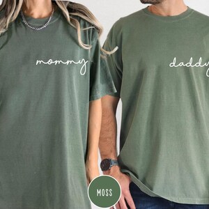 Pu&ograve; includere: Due magliette a maniche corte con girocollo in un colore verde muschio tenue. La maglietta a sinistra presenta la parola mommy in corsivo bianco sul petto, mentre quella a destra presenta la parola daddy in corsivo bianco sul petto.