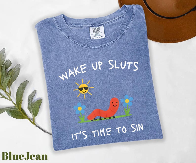 Puede incluir: Camiseta azul con el texto "WAKE UP SLUTS" y "IT'S TIME TO SIN". La camiseta presenta un sol de dibujos animados con gafas de sol, una oruga roja y flores azules. La camiseta es de la marca Comfort Colors.