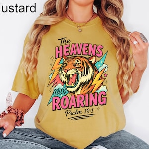 Puede incluir: Camiseta de color mostaza con un gr&aacute;fico de tigre y el texto "The Heavens are Roaring" y "Psalm 19:1". El tigre es naranja, negro y blanco, con acentos rosas y azules. El texto es en rosa y verde.