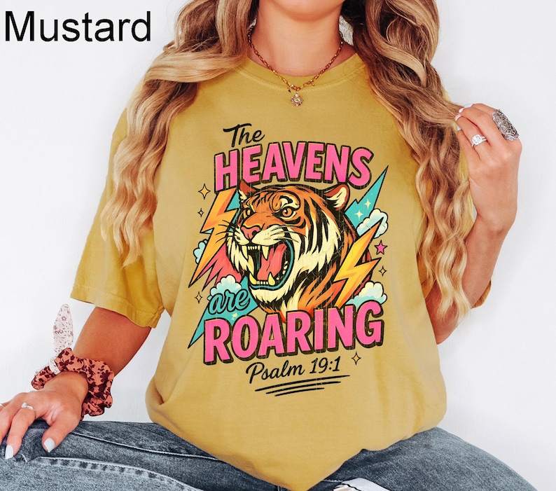 Puede incluir: Camiseta de color mostaza con un gr&aacute;fico de tigre y el texto "The Heavens are Roaring" y "Psalm 19:1". El tigre es naranja, negro y blanco, con acentos rosas y azules. El texto es en rosa y verde.