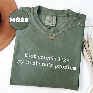 May include: A moss green t-shirt with the text "that sounds like my husband's problem" in white. A gold necklace with a small pendant rests on the shirt. The shirt is folded and displayed with a brown hat and dried plant.