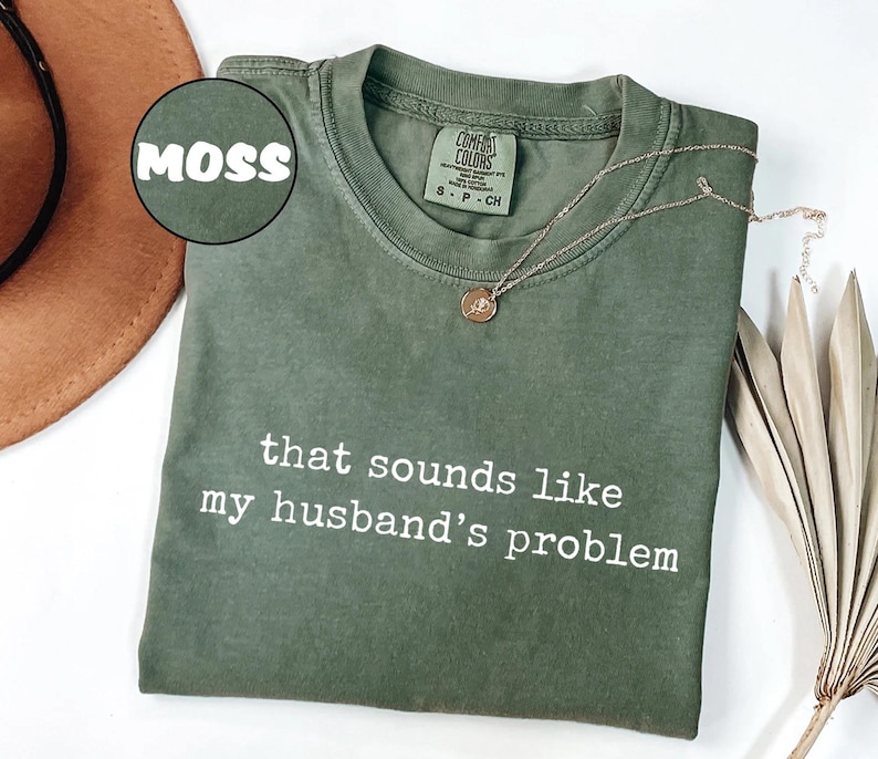 May include: A moss green t-shirt with the text "that sounds like my husband's problem" in white. A gold necklace with a small pendant rests on the shirt. The shirt is folded and displayed with a brown hat and dried plant.