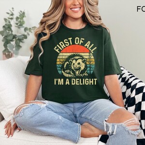 May include: Dark green t-shirt with a distressed graphic of an opossum inside a circle with the text "FIRST OF ALL I'M A DELIGHT". The shirt is a relaxed fit and is made of a soft material.
