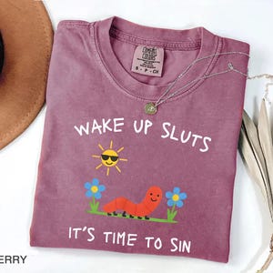 Puede incluir: Camiseta color bayas con el texto "WAKE UP SLUTS IT'S TIME TO SIN". El gr&aacute;fico incluye un sol sonriente con gafas de sol, un gusano rojo y flores azules. Un collar plateado con un colgante est&aacute; sobre la camiseta.
