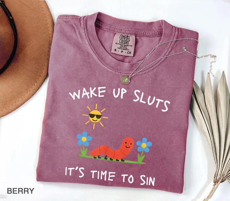 Puede incluir: Camiseta color bayas con el texto "WAKE UP SLUTS IT'S TIME TO SIN". El gr&aacute;fico incluye un sol sonriente con gafas de sol, un gusano rojo y flores azules. Un collar plateado con un colgante est&aacute; sobre la camiseta.