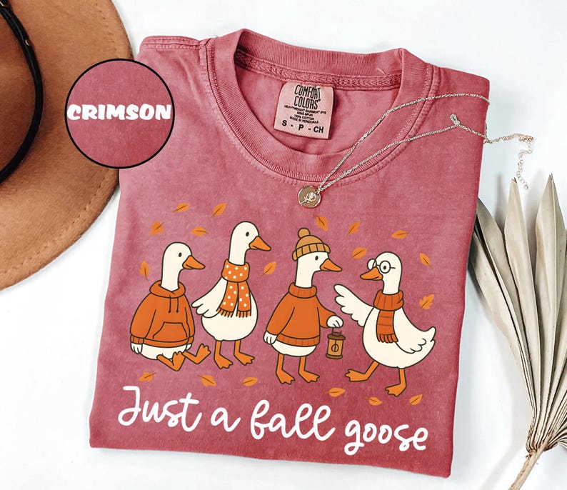 Puede incluir: Camiseta de color carmes&iacute; con una ilustraci&oacute;n de cuatro gansos vestidos con atuendos oto&ntilde;ales. Los gansos llevan su&eacute;teres, bufandas y sombreros, con el texto "Just a fall goose". Un collar plateado est&aacute; drapeado sobre la camiseta.