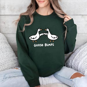 May include: Forest green sweatshirt featuring a cartoon design of two white geese with the text "Goose Bumps" in white. The geese are facing each other with a burst of lines emanating from their beaks. The sweatshirt is a solid color.