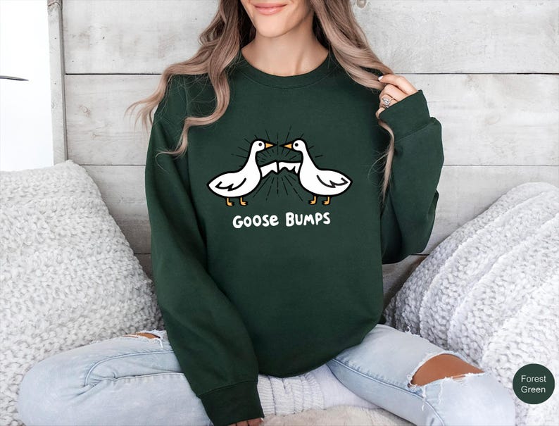 May include: Forest green sweatshirt featuring a cartoon design of two white geese with the text "Goose Bumps" in white. The geese are facing each other with a burst of lines emanating from their beaks. The sweatshirt is a solid color.