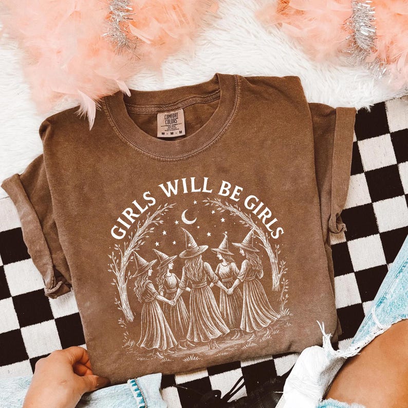 May include: Brown t-shirt with the text "GIRLS WILL BE GIRLS" in white. The graphic features five figures in witch hats holding hands under a crescent moon and stars. The shirt is a Comfort Colors brand.